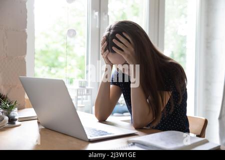 Donna sconvolto che stringe la testa sedersi alla scrivania con un computer portatile Foto Stock