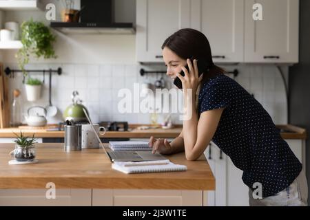 La donna parla di cellulare uso laptop in cucina Foto Stock