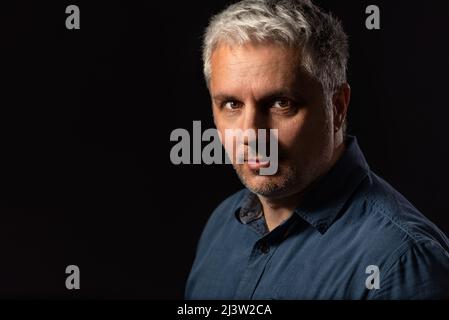 Testa e spalle basso ritratto chiave di un uomo bearded di mezza età guardando attentamente la fotocamera su uno sfondo nero studio con spazio copia Foto Stock