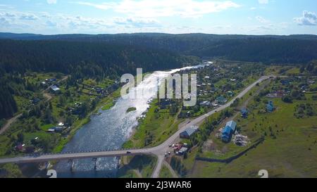 Vista dall'alto. Clip. Una bella foresta grande vicino a piccole case e una zona residenziale e un grande fiume lungo e un cielo luminoso dall'alto. Foto Stock