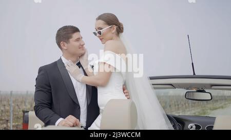 Appena sposato coppia, sposa e sposo all'aria aperta. Azione. Bella donna in abito bianco e occhiali da sole che abbracciano l'uomo in vestito durante la fotosessione Foto Stock