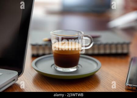 Espresso fresco in una tazza di caffè di vetro su una scrivania in legno vista ravvicinata. Computer portatile e spazio di lavoro aziendale Foto Stock