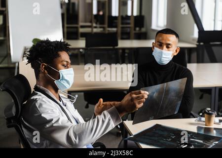 Paziente asiatico alla nomina del medico africano. Frattura delle ossa del piede. Chirurgo nero che mostra la radiografia al paziente Foto Stock