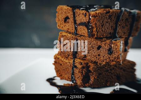 Scatto molto vicino di un pile di brownie al cioccolato con ciocche di cioccolato che cadono su un piatto bianco e sfondo scuro. Vista normale. Foto Stock