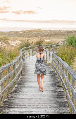 Donna bionda che cammina su passerella in legno o ponte in abito alla moda soffiato dal vento Foto Stock