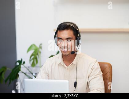 Un uomo asiatico sorride indossando una cuffia che guarda il suo laptop in ufficio Foto Stock
