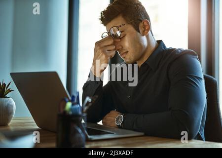 Quando lo stress del lavoro innesca emicranie gravi. Scatto di un giovane uomo d'affari che guarda stressato mentre lavora su un notebook in ufficio. Foto Stock