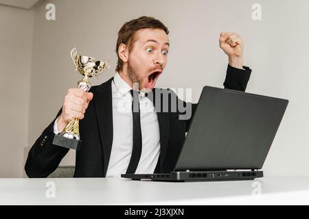 Un uomo d'affari di successo in tuta che celebra la vittoria con il trofeo della coppa d'oro mentre guarda la cerimonia online sul laptop. Foto Stock