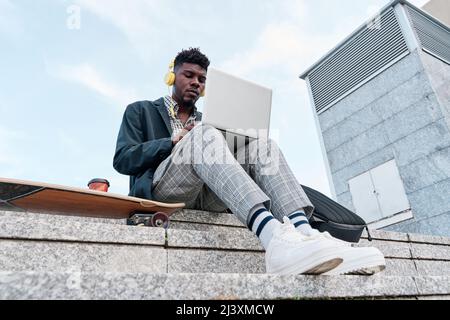 Vista dal basso di un giovane adulto afroamericano su uno skateboard mentre lavora su un computer portatile Foto Stock