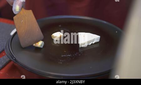 Friggere il formaggio a mani ravvicinate in una padella. ARTE. Il cuoco prepara il formaggio alla fiera. Cuoco gira su tofu con una spatola di legno Foto Stock