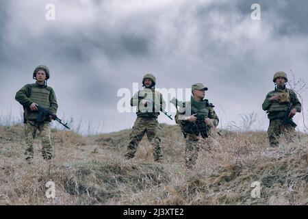 Squadra di quattro soldati ucraini si erge con fucili d'assalto kalashnikov tra erba secca sullo sfondo del cielo e delle nuvole. Concetto di milita russa Foto Stock