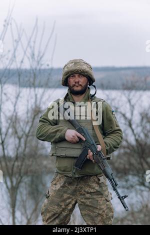 Il soldato ucraino è in piedi con il fucile d'assalto kalashnikov nelle sue mani sullo sfondo del cielo e del fiume. Concetto di invasione militare russa in Ukra Foto Stock