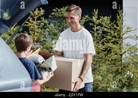 Ritratto di sorridente padre e figlio che porta scatole in auto mentre si sposta in casa nuova illuminata dalla luce del sole all'aperto Foto Stock
