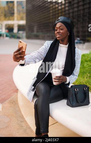 Sorridente femmina afroamericana che indossa abiti casual e un foulard musulmano seduto con caffè da asporto e che prende autoritratto sul cellulare in città Foto Stock