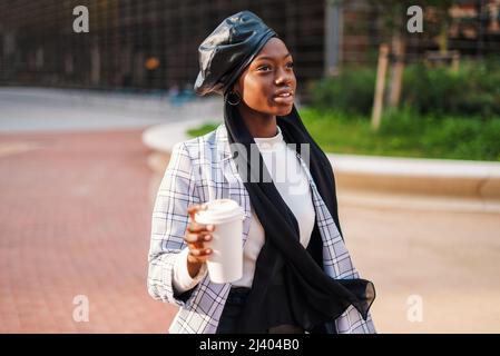 Seria giovane afroamericana donna in abito trendy con berretto in pelle sul foulard musulmano che ha takeaway bevanda e guardare via con attento sguardo nel parco Foto Stock