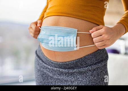 Gravidanza durante la pandemia di coronavirus. Donna incinta che copre il ventre con maschera facciale per il concetto di prevenzione del virus corona. Le donne in gravidanza sono più alte Foto Stock