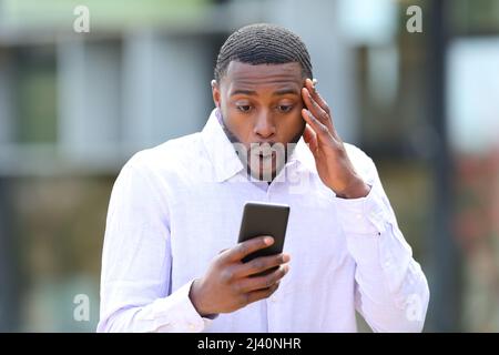 Vista frontale ritratto di un uomo sorpreso con pelle nera leggere notizie al telefono in strada Foto Stock