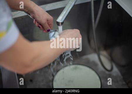 La donna lava i piatti sotto flusso d'acqua. Dettagli cucina. La lavastoviglie pulisce il cucchiaio nel lavandino. L'acqua calda di fondo si riversa dal rubinetto. Foto Stock