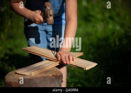 Il bambino indurisce il chiodo in asse. La ragazza impara a battere con il martello. Il preschooler è inserito nella manodopera manuale. Apprendimento senza computer. Lavorare all'aria aperta. Foto Stock