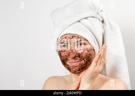 Una bella giovane donna con un asciugamano da bagno sulla testa e una maschera di caffè sul viso si prende cura della sua pelle Foto Stock