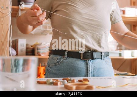 Le mani della donna croppate che tengono e stringano i branelli. Giocattolo ecologico in legno con decorazione a chiazze di trinket sulla scrivania bianca. Lavori di artigianato in negozio Foto Stock