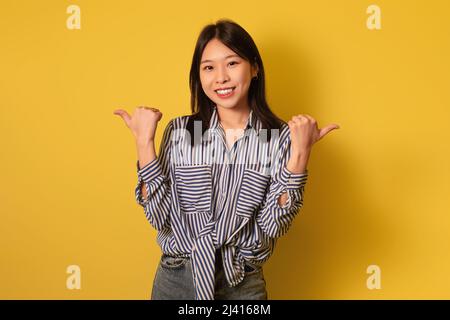 Gioiosa giovane donna asiatica in abbigliamento casual mostrando i pollici in su con entrambe le mani, sorridendo alla macchina fotografica su sfondo giallo Foto Stock