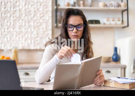 Donna concentrata leggere interessante libro di carta mentre si siede al tavolo in cucina interno, trascorrere il tempo libero a casa Foto Stock