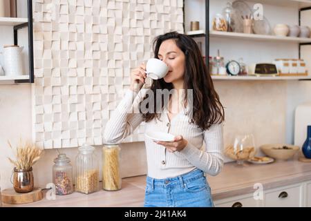 Donna allegra in abiti casual bevendo la sua bevanda calda preferita in tazza bianca, in piedi in cucina moderna interno Foto Stock