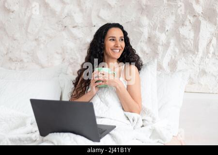 Allegra giovane ragazza bruna europea attraente con capelli lunghi drink caffè da tazza, si siede a letto con un computer portatile Foto Stock