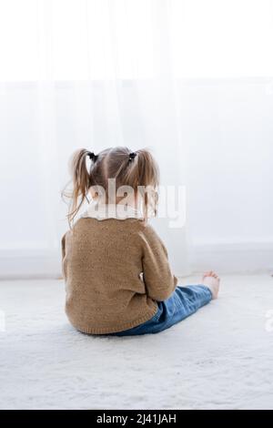 vista posteriore della bambina offesa con ponytail seduta sul pavimento a casa Foto Stock
