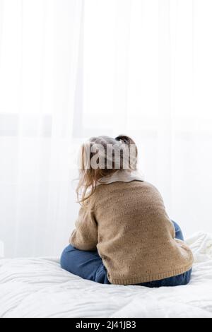 vista posteriore della bambina con code di pony seduta da sola sul letto di casa Foto Stock