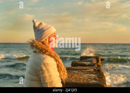 Una donna con capelli biondi in un cappello e un cappotto di pelliccia cammina lungo il mare lungo una pietra frangiflutti Foto Stock