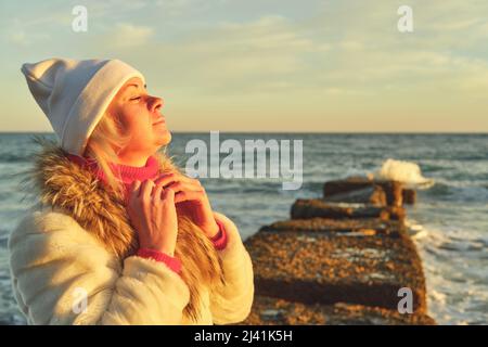 Una donna con capelli biondi in un cappello e un cappotto di pelliccia cammina lungo il mare lungo una pietra frangiflutti Foto Stock
