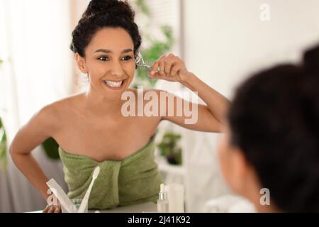 Bella giovane donna utilizzando ciglia arricciatore di fronte allo specchio a casa, spazio libero Foto Stock