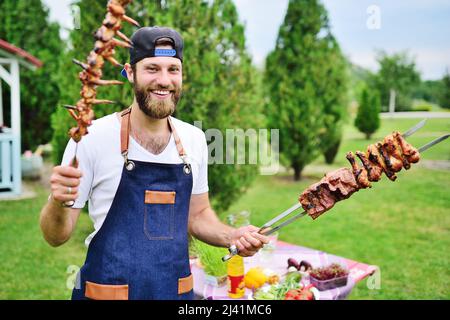 il giovane uomo portico in un cappellino e grembiule di denim sorride e tiene uno spiedino con deliziosa carne alla griglia cucinata sullo sfondo di Foto Stock