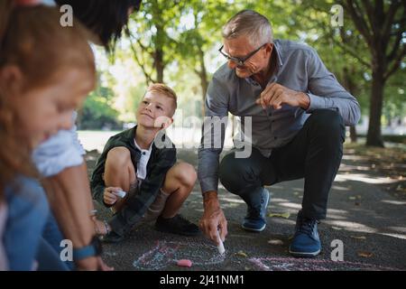 Coppia senior con nipoti che disegnano con gessi su marciapiedi all'aperto nel parco. Foto Stock