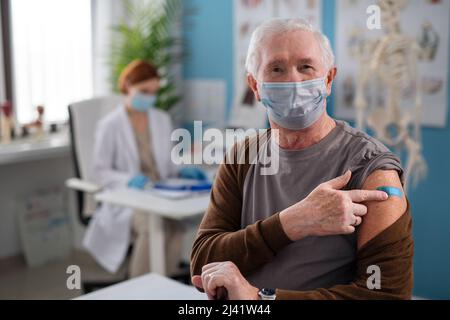Uomo anziano che mostra bendaggio dopo essere stato vaccinato nell'ufficio del medico, guardando la macchina fotografica. Foto Stock