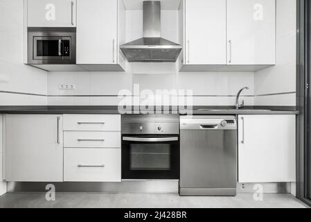 Vista frontale della cucina con mobili in legno bianco con ripiani grigi, elettrodomestici in acciaio inox e pavimenti piastrellati. Foto Stock