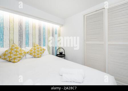 Camera da letto con letto matrimoniale con biancheria bianca, cuscini a motivi gialli, pareti decorate con carta da parati e guardaroba a porte scorrevoli bianche in stile veneziano Foto Stock