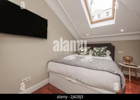 Camera da letto con letto matrimoniale con cuscini e coperte in denim, cuscini verdi, lucernario nel soffitto, tv appeso alla parete, asciugamani bianchi nella mansarda Foto Stock