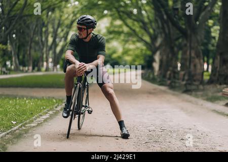 Giovane sportivo che si diverta con l'hobby attivo preferito al parco verde della città. Ciclista stanco in casco e occhiali seduti in bici nera, prendendo pausa e lookin Foto Stock