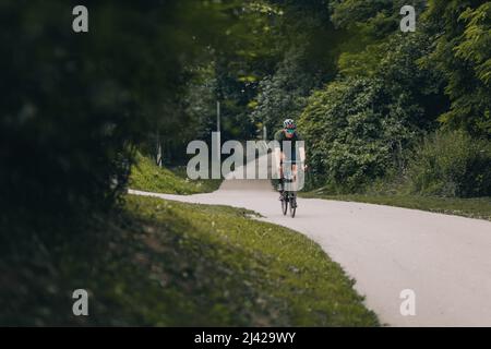 Sportivo caucasico in abbigliamento sportivo, casco protettivo e occhiali a specchio in bicicletta presso il parco cittadino. Uomo forte che fa il mattino di allenamento all'aria fresca. Foto Stock