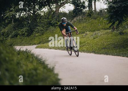 Giovane uomo con gambe muscolari che indossa casco di sicurezza e occhiali da corsa da solo nel verde parco cittadino. Formazione professionale di ciclista regolarmente su aria fresca. Foto Stock