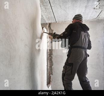 Processo di decostruzione del divisorio dell'intercapedine in appartamento. Vista laterale del costruttore maschio che lavora con martello pneumatico all'interno dell'appartamento. Foto Stock