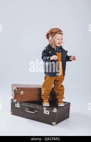 Un ragazzino in giacca di pelle e tute marroni si trova su una grande valigia. Bambino con una valigia. Piccolo viaggiatore. Foto Stock