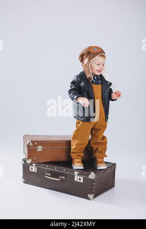 Un ragazzino in giacca di pelle e tute marroni si trova su una grande valigia. Bambino con una valigia. Piccolo viaggiatore. Foto Stock