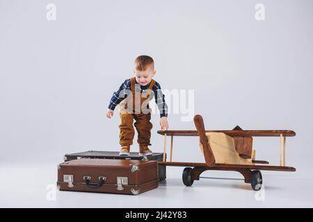 Un bambino allegro in una tuta marrone è in piedi su una valigia. Piccolo viaggiatore con una valigia. Aereo in legno, giocattoli ecologici per bambini Foto Stock