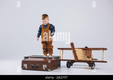 Un bambino allegro in una tuta marrone è in piedi su una valigia. Piccolo viaggiatore con una valigia. Aereo in legno, giocattoli ecologici per bambini Foto Stock