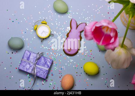 Elegante composizione pasquale - uova colorate multicolore dipinte, sveglia retrò gialla, scatola regalo lilla, biscotti a forma di un top viola di Pasqua coniglietto Foto Stock