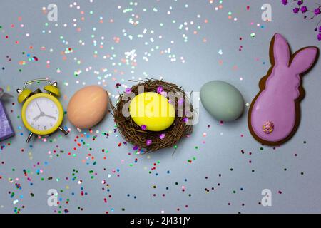 Composizione di Pasqua - un nido con uova dipinte di giallo e blu, sveglia retrò, scatola regalo lilla, biscotti di pan di zenzero a forma di una Pasqua viola Foto Stock